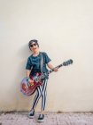 Дерзкий активный возбужденный веселый мальчик в красочной одежде, играющий на гитаре на фоне белой стены — стоковое фото