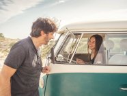 Vue latérale de type souriant homme parlant à jolie femme riant sur le siège avant de la voiture dans un endroit désert — Photo de stock