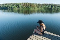 Side view of young woman in black swimsuit and hat sitting on wooden pier on the mobile phone on a lake on clear blue sky and forest background — Stock Photo