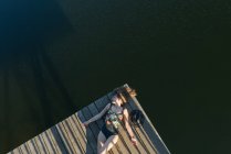 Top view of beautiful woman in black swimsuit and hat lying down on wooden pier of lake on clear blue sky and forest background — Stock Photo