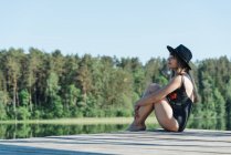 Vista laterale di felice giovane donna in costume da bagno nero e cappello seduto sul molo di legno e ammirando la vista del lago su cielo blu chiaro e sfondo della foresta — Foto stock