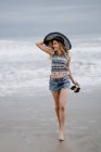 Attractive femme en chapeau noir tenant sac de plage et chaussures tout en profitant d'une vue pittoresque sur l'océan regardant loin — Photo de stock
