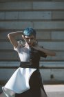 Jeune femme en robe futuriste touchant le visage et les cheveux bleus courts tout en se tenant près du bâtiment par une journée ensoleillée sur la rue de la ville — Photo de stock
