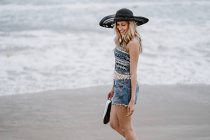 Приваблива жінка в чорному капелюсі тримає пляжний мішок і взуття, насолоджуючись мальовничим видом на океан, дивлячись вниз — стокове фото