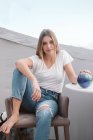 Sorridente donna casual in jeans strappati e t-shirt bianca rilassante in sedia e guardando in macchina fotografica — Foto stock