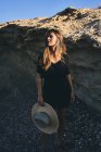 Joven modelo femenina sonriente de pelo largo mirando hacia otro lado sosteniendo el sombrero en la costa rocosa - foto de stock