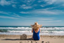 Vista posterior de la mujer bonita en sombrero y traje de baño sentado con bolsa en la playa de arena mirando las olas bajo el cielo turquesa nublado - foto de stock