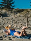 Vista posterior de la mujer en sombrero y traje de baño azul libro de lectura mientras se encuentra en la playa de arena en el día soleado - foto de stock