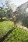 Pequeno garoto rindo em shorts e com os pés descalços salpicando água para a câmera da mangueira de jardim — Fotografia de Stock