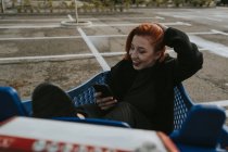 Улыбающаяся женщина со смартфоном в тележке для покупок на парковке — стоковое фото