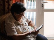 Пожилая седовласая женщина в рубашке и очках сидит на кресле и читает электронную книгу в квартире — стоковое фото