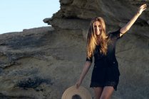 Молодая улыбающаяся женщина смотрит в камеру, держа шляпу на скалистом побережье — стоковое фото