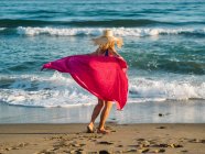 Радостная женщина в шляпе и голубом купальнике, размахивающая красным шарфом и танцующая вдоль моря в солнечный светлый день — стоковое фото