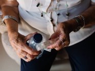Imagen recortada de mujer mayor en camisa blanca sosteniendo píldoras y agua de la botella, sentado en el sillón en el apartamento - foto de stock