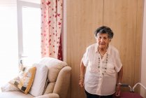 Anziana donna in camicia bianca con mano sulla vita in piedi mentre appoggiata sulla poltrona a casa e guardando la fotocamera — Foto stock