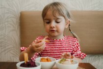 Cute little girl eating assorted tasting appetizing snack at table — Stock Photo