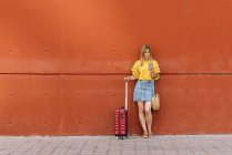 Молода жінка-туристка використовує мобільний телефон, спираючись на червону стіну — стокове фото