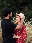 Mann umarmt lächelnde schwangere Frau, während er Strohhut vor dem Hintergrund des malerischen grünen Parks justiert — Stockfoto