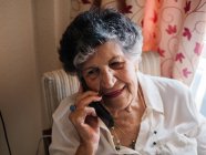 Heureuse femme âgée souriante et parlant sur un téléphone portable tout en regardant loin à la maison — Photo de stock