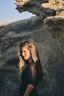 Junge langhaarige stilvolle nachdenkliche Frau steht im Sonnenlicht mit Felsen auf dem Hintergrund — Stockfoto