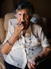 Элегантная пожилая женщина, принимающая таблетки, сидящая дома на кресле и смотрящая в камеру — стоковое фото