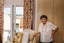 Anziana donna in camicia bianca con mano sulla vita in piedi mentre appoggiata sulla poltrona a casa e guardando la fotocamera — Foto stock