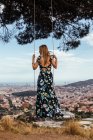 Дівчина на спині у квітковій сукні, що розмірковує про місто під час сходження на гойдалку — стокове фото