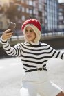 Young smiling woman in French red cap, striped blouse and white shorts taking photo on urban background — Stock Photo