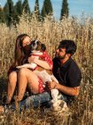 Улыбающаяся веселая пара, сидящая среди высокой травы и развлекающаяся с маленькой собачкой в сельской местности — стоковое фото