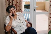 Счастливая пожилая женщина улыбается и разговаривает по мобильному телефону, отводя взгляд от дома — стоковое фото