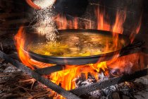 Aggiunta di riso in una grande padella di ferro con brodo bollente per cuocere la paella sul fuoco aperto con la legna — Foto stock