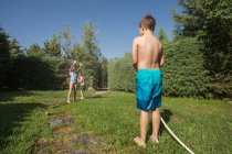 Crianças em trajes de banho correndo e salpicando água da mangueira de jardim umas para as outras — Fotografia de Stock