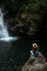 Веселая женщина-путешественница в шляпе улыбается и смотрит в камеру, сидя на мокром валуне возле водопада — стоковое фото
