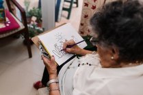 From above of senior woman in white shirt and glasses drawing on paper with pencil, sitting on armchair at home — Stock Photo