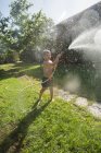 Kleines lachendes Kind in kurzen Hosen und mit nackten Füßen, die Wasser aus Gartenschlauch spritzen — Stockfoto
