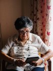 Senior femme en chemise et lunettes assis sur le fauteuil et la lecture e-book dans l'appartement — Photo de stock