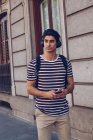 Привлекательный молодой человек в повседневной одежде слушает музыку во время прогулки по улице при свете дня — стоковое фото