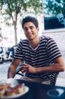Portrait de jeune homme joyeux en t-shirt dépouillé regardant à la caméra alors qu'il était assis sur une terrasse tenant un verre de bière — Photo de stock