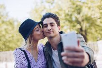Joven hombre guapo tomando selfie con novia mientras se besa en hermoso jardín - foto de stock