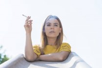 Молода блондинка курить сигарету і дивиться на камеру на білому тлі, спираючись на поручні сходів — стокове фото