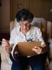 Femme âgée en chemisier et lunettes assis sur un fauteuil près de la fenêtre et la résolution de mots croisés à la maison — Photo de stock