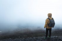 Back view of guy with backpack standing on hillside against thick fog during trip in nature — Stock Photo