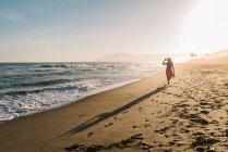 Visão traseira da mulher magra em chapéu e pareo andando ao longo da costa em luz solar brilhante — Fotografia de Stock