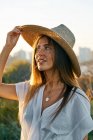 Молода усміхнена жінка в білому одязі і капелюсі дивиться в природу на заході сонця — стокове фото