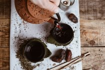 Mão pessoa irreconhecível servindo chá saboroso fragrante em xícara bule de barro e datas doces na bandeja branca decorada com folhas de chá no fundo de madeira — Fotografia de Stock