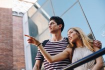 Привлекательная молодая пара туристов, любующихся видом и указывающих, стоя перед современным зданием — стоковое фото