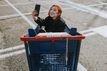 Smiling woman taking selfie with smartphone in shopping trolley in parking lot — Stock Photo