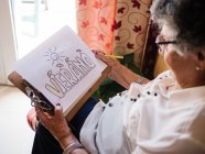 Senior femme aux cheveux gris en chemise blanche et lunettes dessin sur papier avec crayon, assise sur fauteuil à la maison — Photo de stock