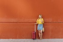 Jovem turista com mala falando no celular enquanto se inclina na parede vermelha — Fotografia de Stock