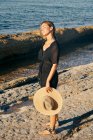 Молода приваблива жінка насолоджується сонцем з закритими очима на пляжі і тримає капелюх — стокове фото